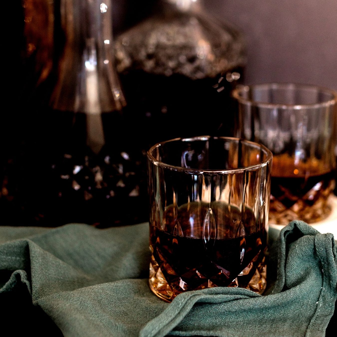 Two crystal whiskey glasses with dark amber liquid on a green cloth with a decanter in the background.