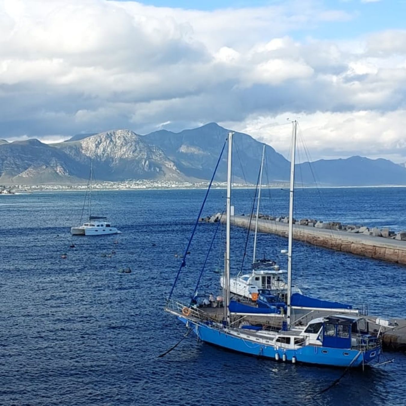 Blue sailboat anchored by a rocky pier with mountains and a town under cloudy skies in the background.