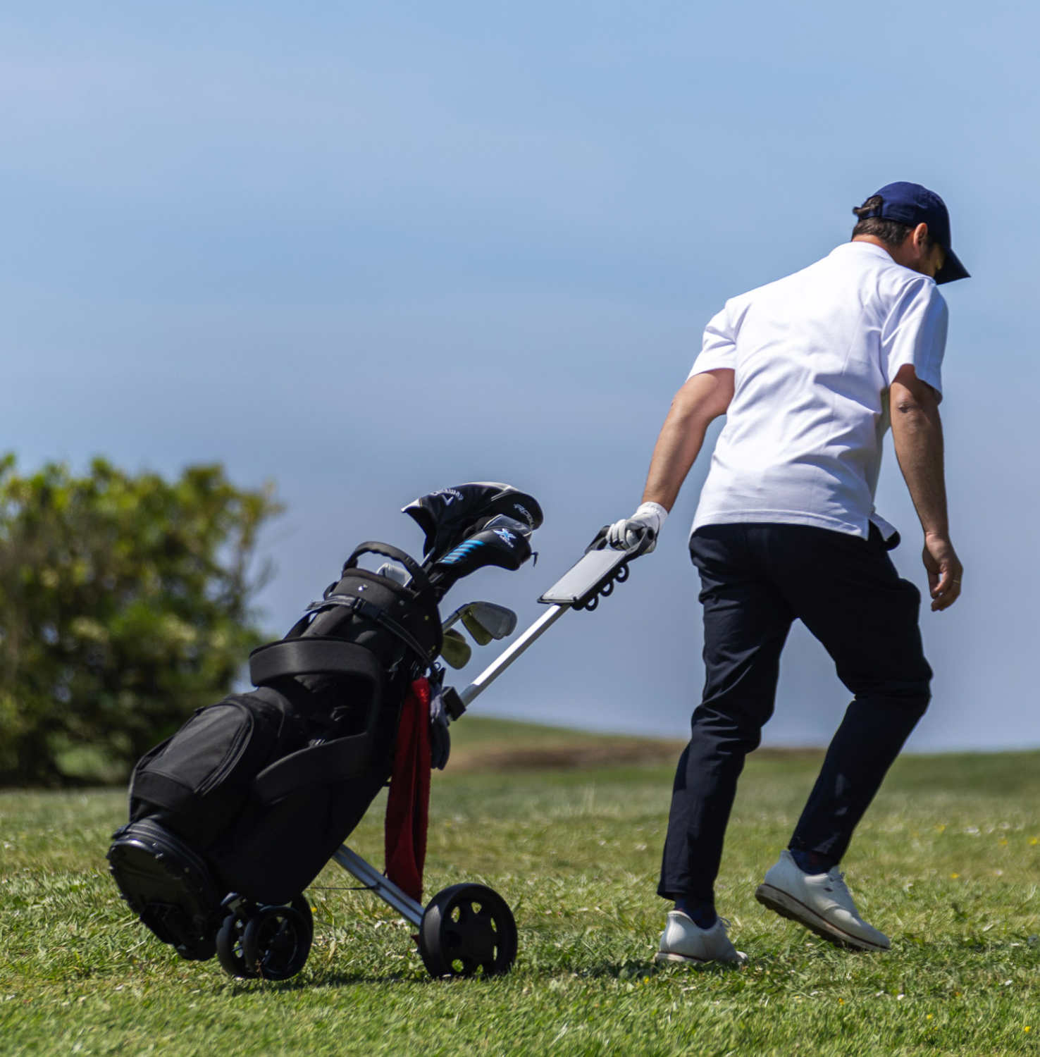 Golfer in white shirt and cap walking on grass, pulling a golf bag trolley with clubs.