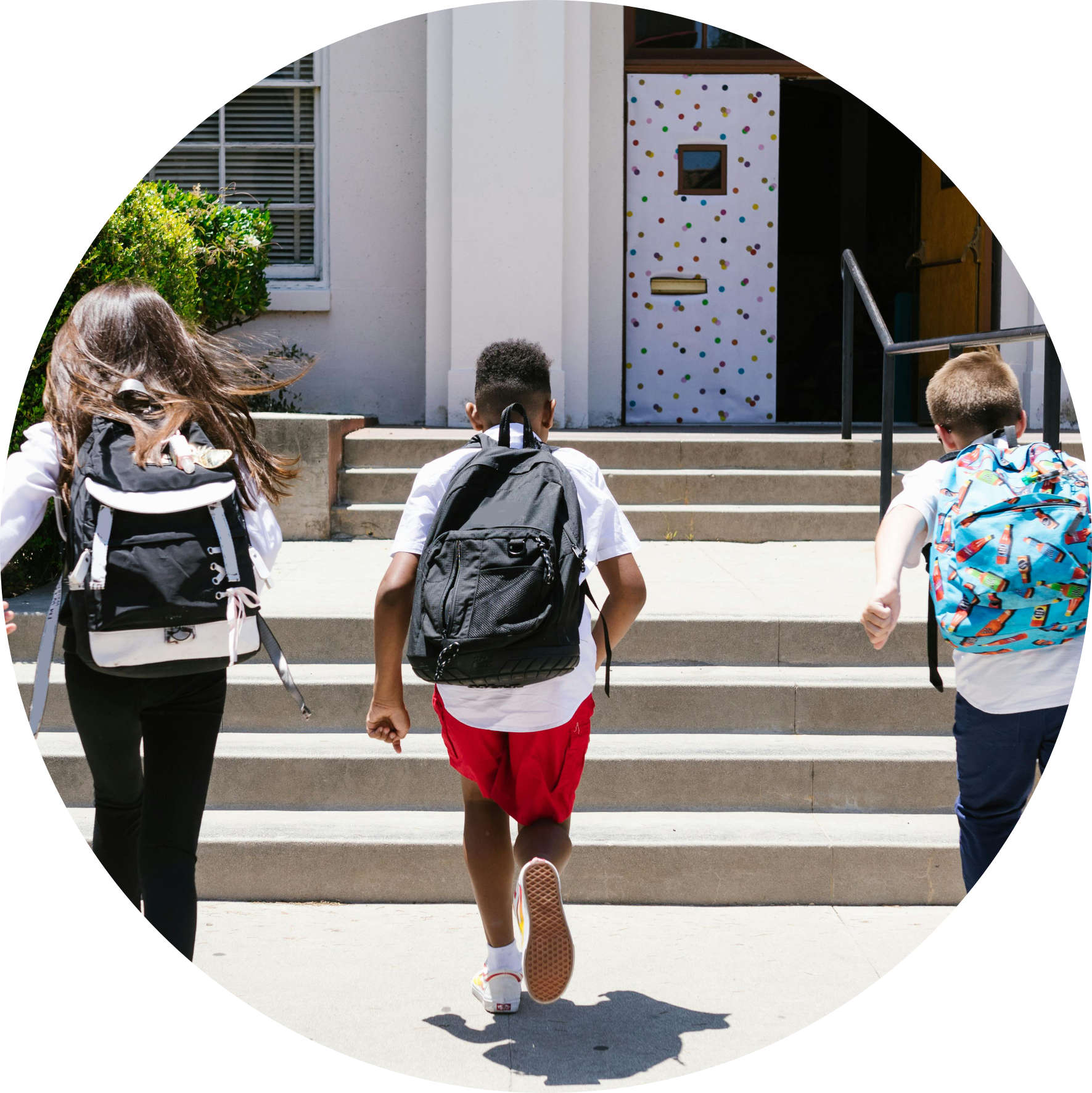 Three children wearing backpacks running up school steps toward a polka-dotted door.
