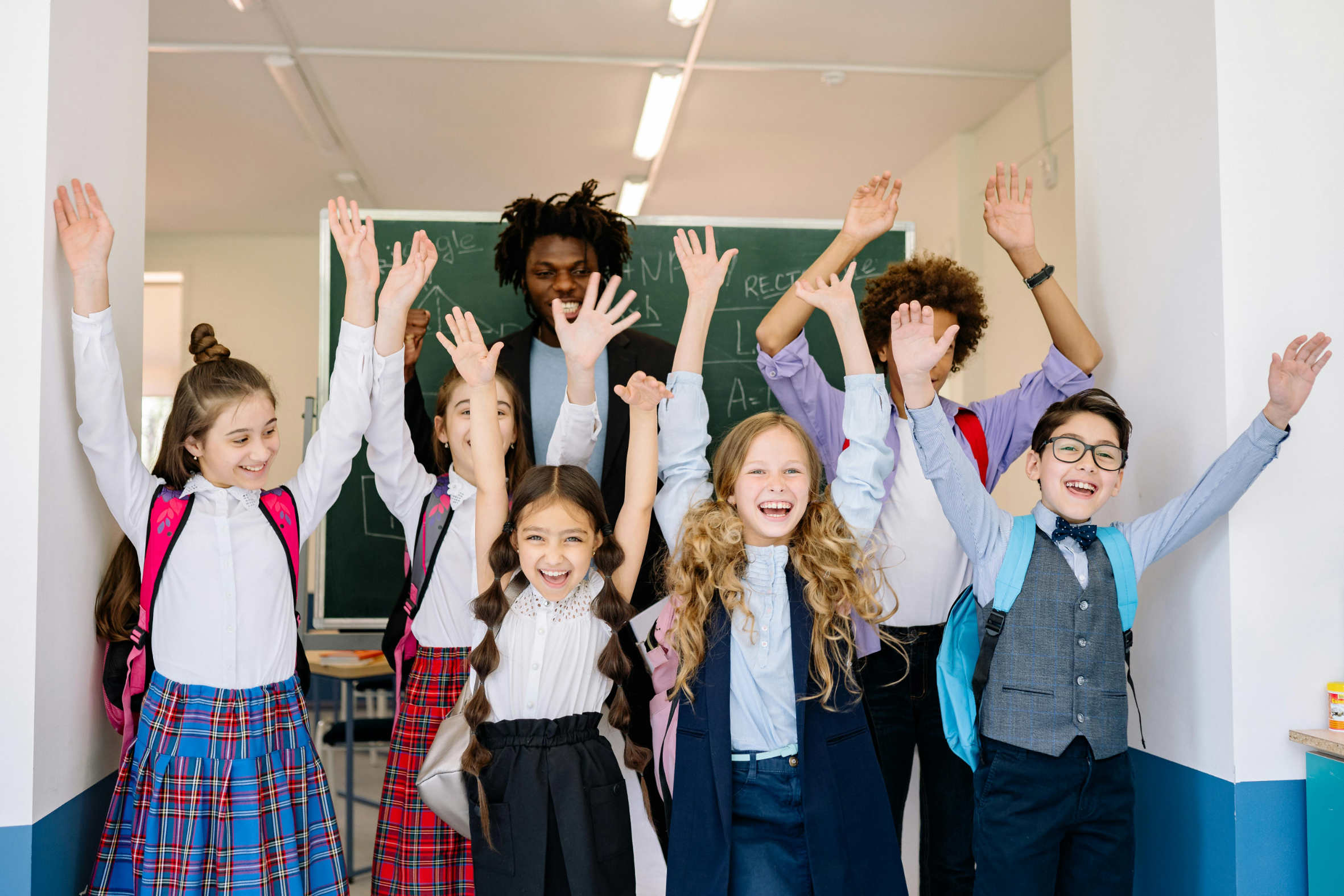 Happy diverse schoolchildren with backpacks raising hands inside classroom near chalkboard.